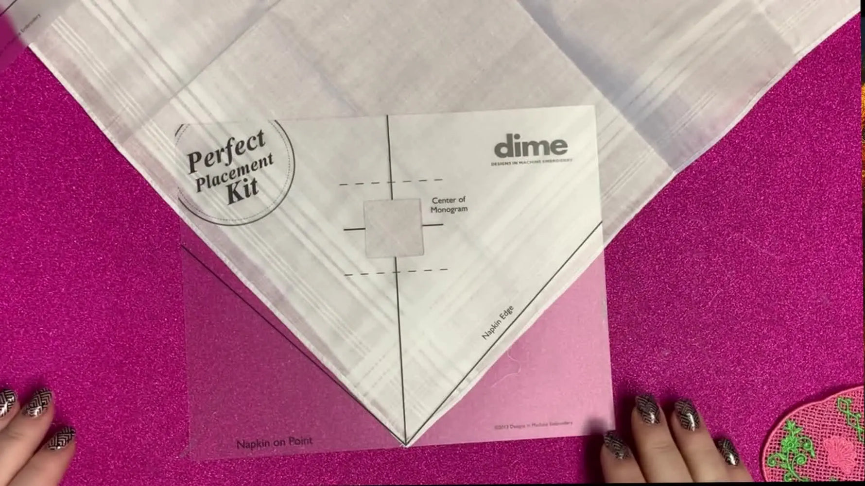 A translucent placement template is aligned over the corner of a white handkerchief, showing a fast, accurate way to find the embroidery center point.