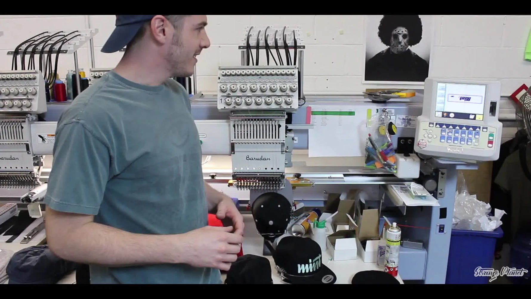 Man in a t-shirt and backward cap, gesturing in front of multiple embroidery machines.