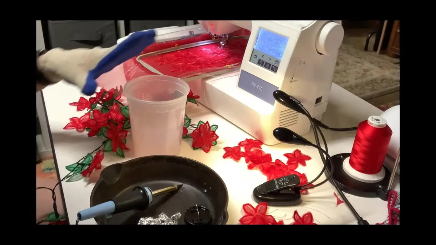 A close-up shows a heat tool sealing the organza edge around a stitched poinsettia piece on a metal pan while a heat-resistant glove protects the hand.