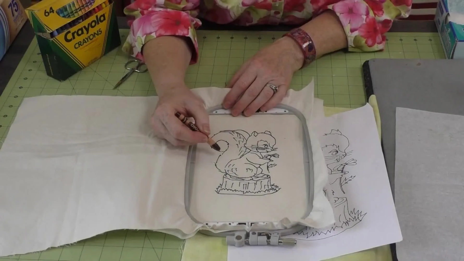 An overhead view of a hand coloring an embroidered squirrel outline on fabric with a brown crayon before heat-setting.