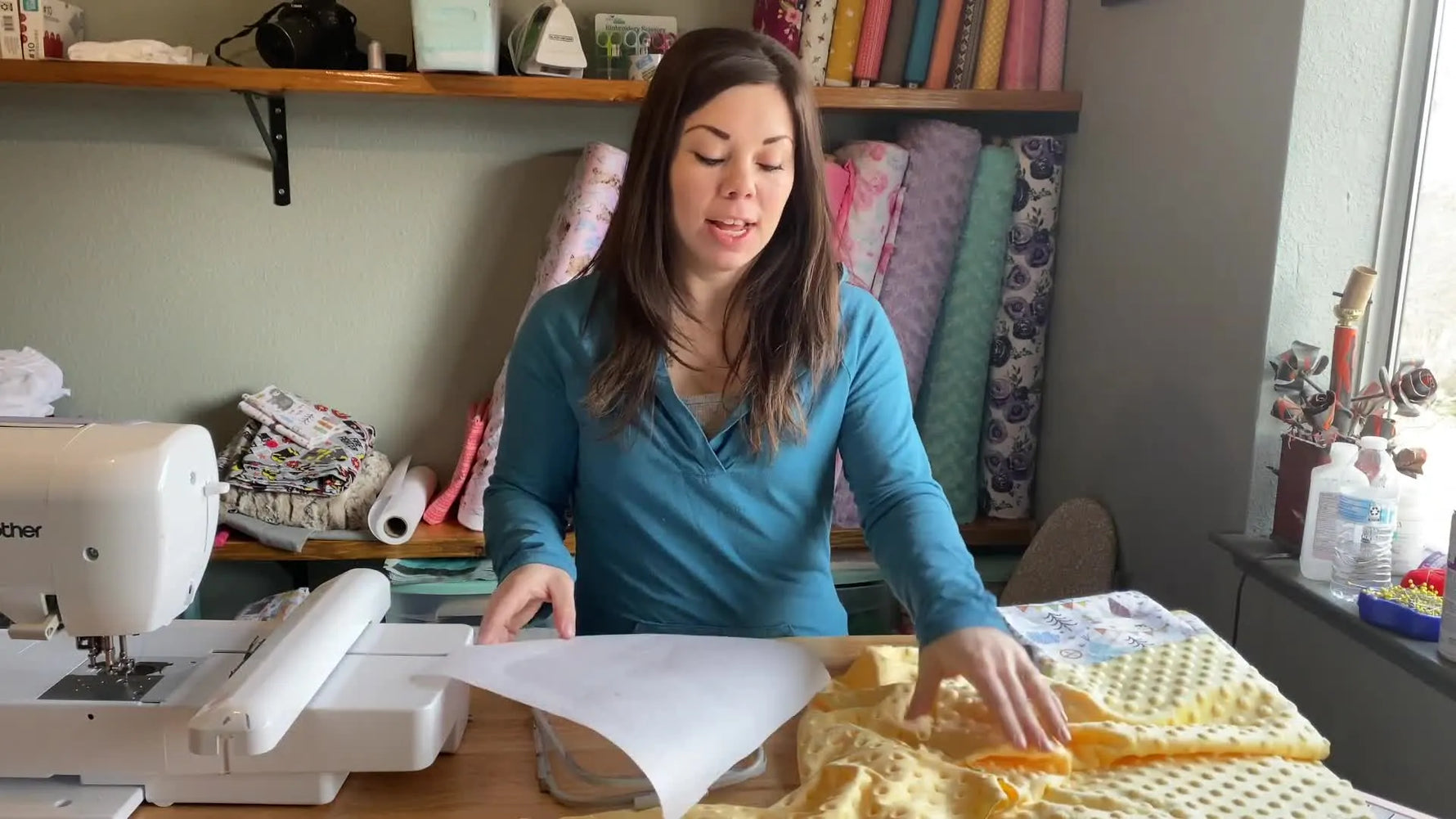 Presenter holding a piece of minky fabric and a roll of stabilizer, with a hoop and basting spray on the table.