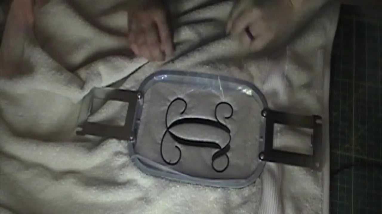 A plush towel sits hooped with a finished monogram “D” visible under a clear heavyweight water-soluble topping film.