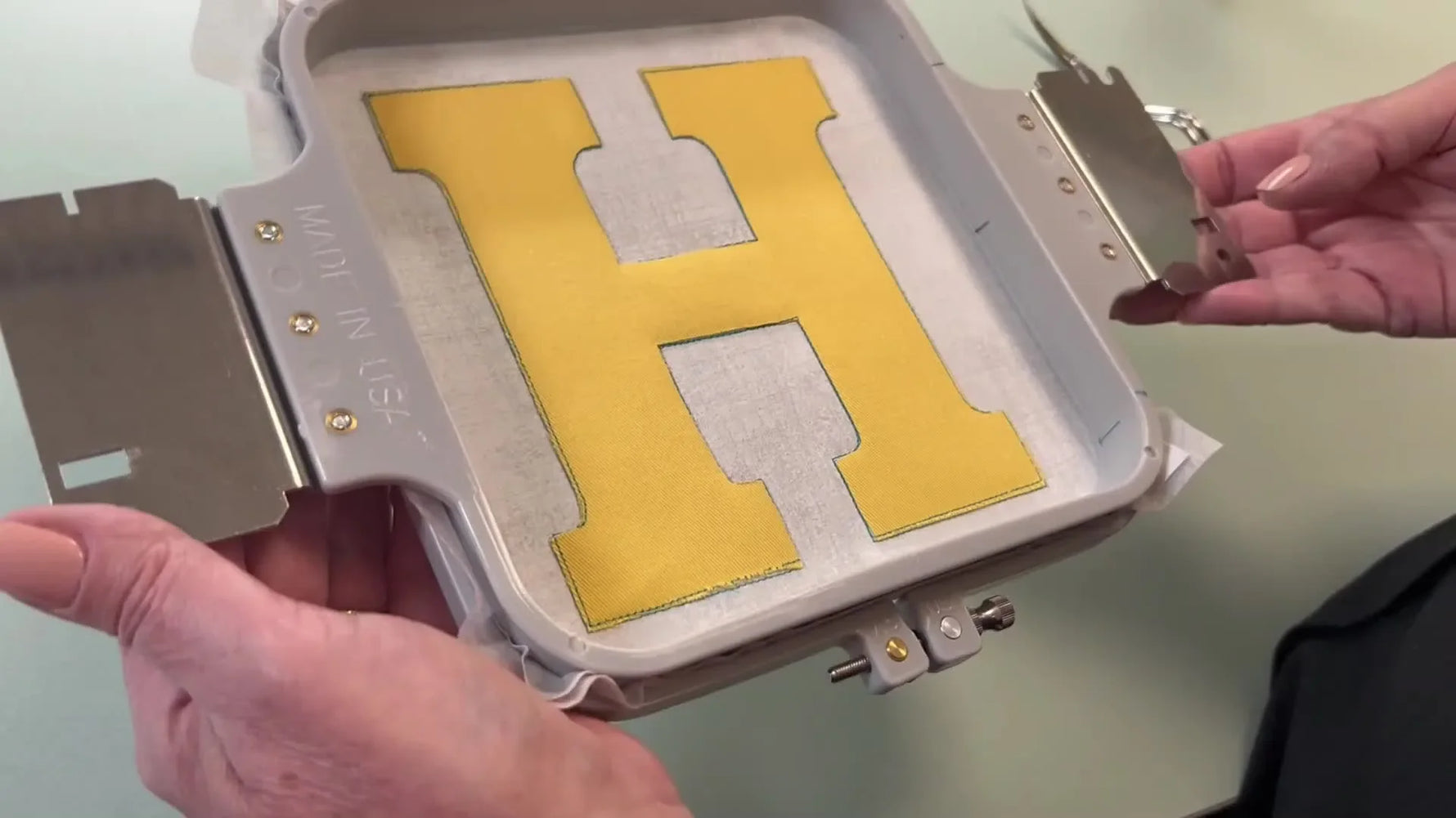 A Tajima machine finishes a wide teal satin stitch border around a yellow appliqué letter H on a quilt block.
