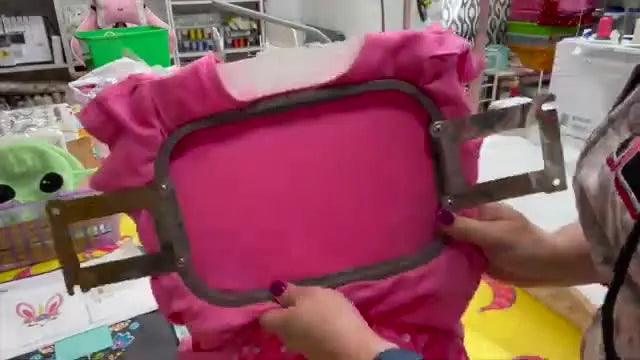 A hooped pink toddler dress is held up to show the stabilizer layers and the standard hoop’s screw mechanism after successful hooping.