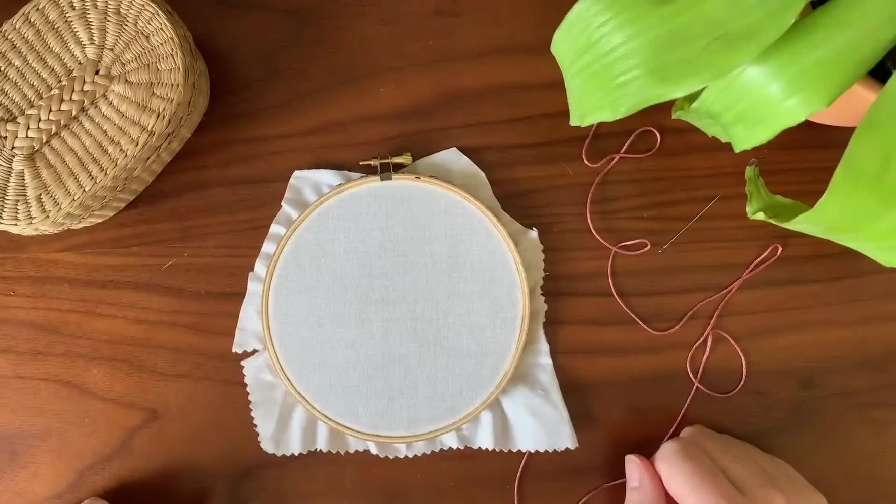 Overhead shot of an embroidery hoop with white fabric on a wooden table, surrounded by crafting supplies.