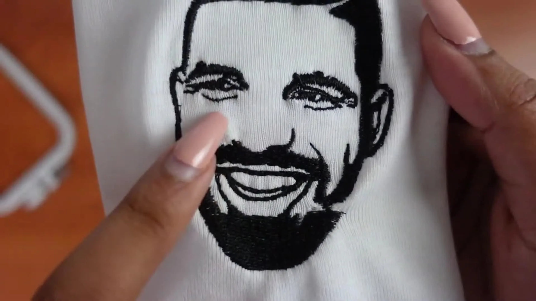 Close-up of a completed black and white embroidered Drake face on white fabric, held by hands with manicured nails.