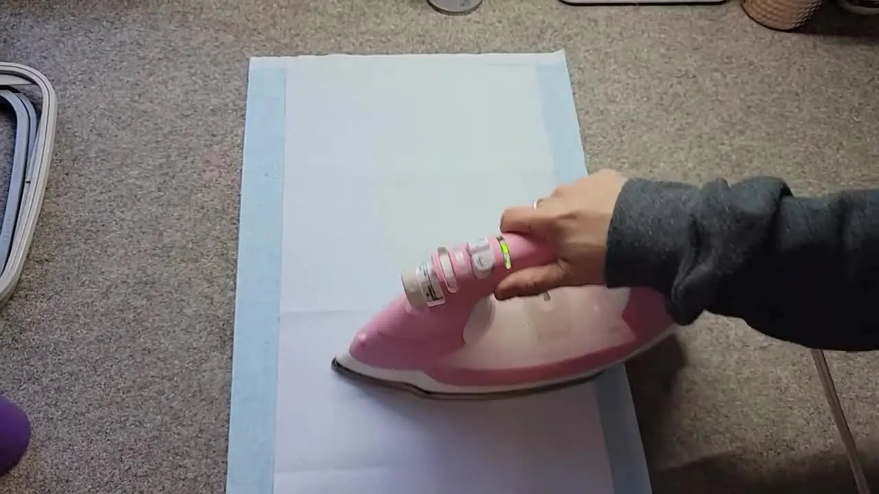 A hot iron fuses a centered strip of Shape Flex interfacing onto a blue cotton background strip to stabilize dense tiling-scene embroidery.