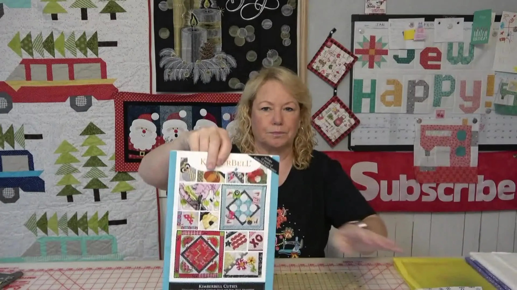 The host holds up the Kimberbell Cuties pattern book cover featuring multiple table topper designs.