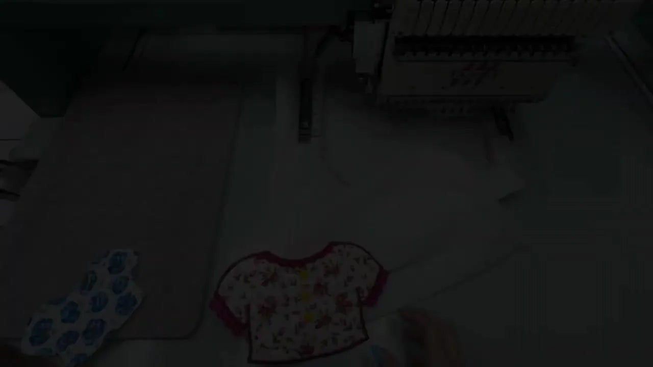A finished floral ITH doll shirt is held up close, showing the lining, lace detail, and snap placement markings.