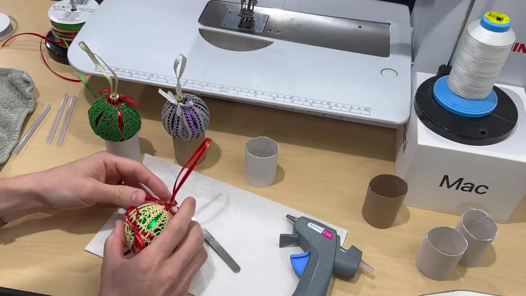 Three finished freestanding lace Christmas ornaments in multiple colorways are displayed upright on paper towel roll stands beside the embroidery machine.