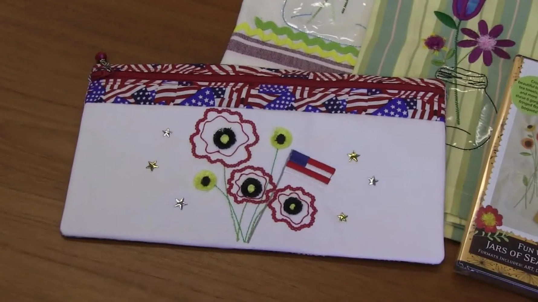 A close-up of a finished white zipper pouch featuring poppy flowers, an American flag motif, and gold star embellishments.