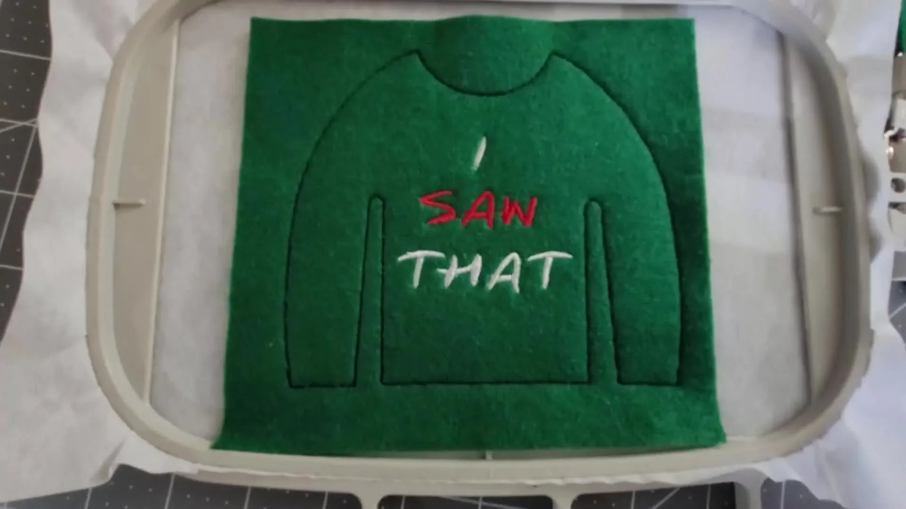 A close-up of the finished “I SAW THAT” embroidery stitched in red and white on green felt inside a 5x7 hoop.