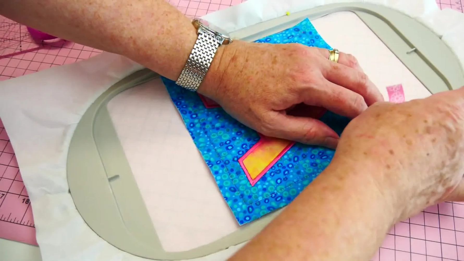A close-up shows pink tape securing a quilt block corner while it’s floated and aligned over a stitched registration outline on tearaway stabilizer.