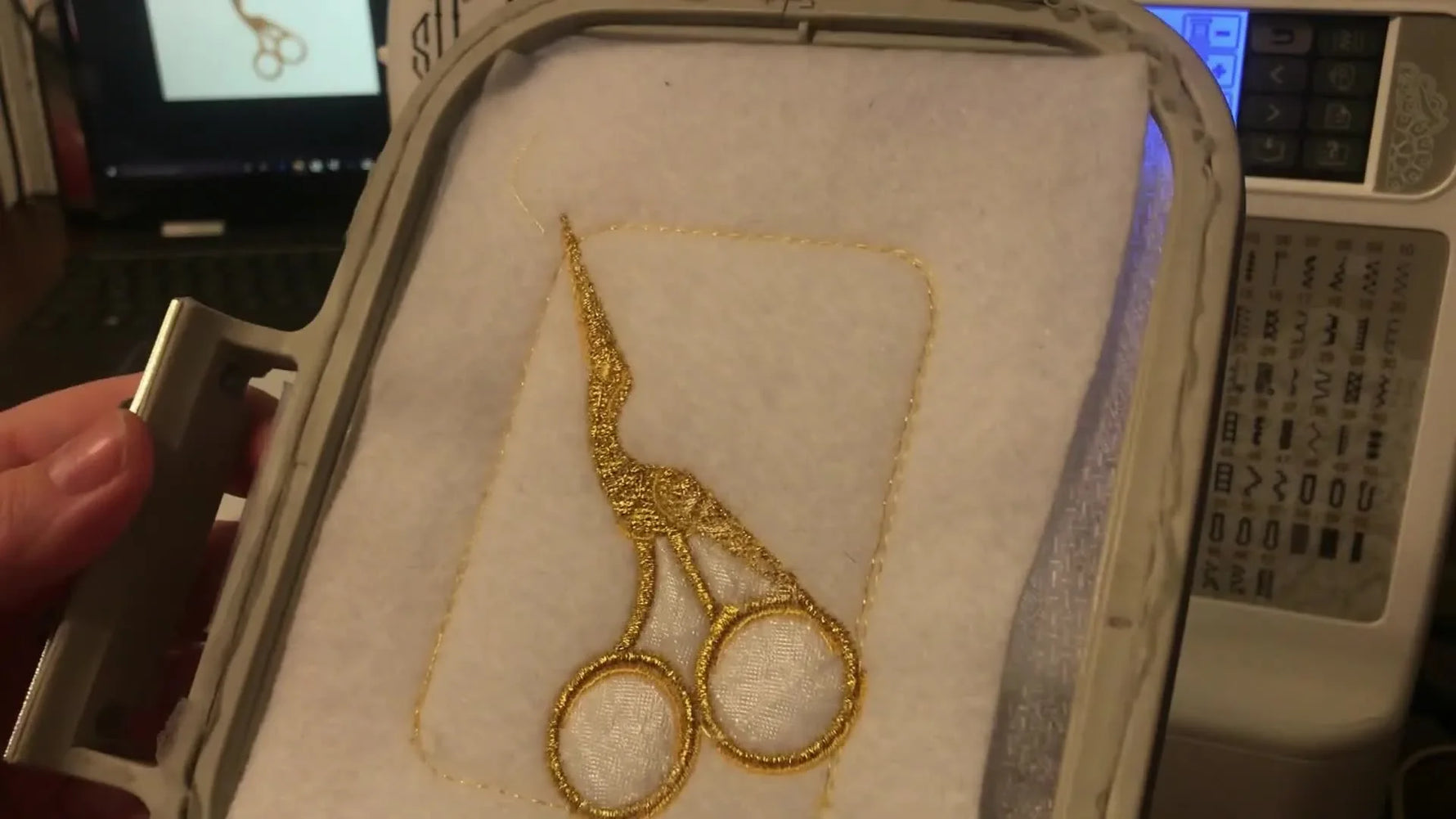 A finished embroidered scissor pouch is shown still in the hoop under the machine light, highlighting the clean border and centered design.