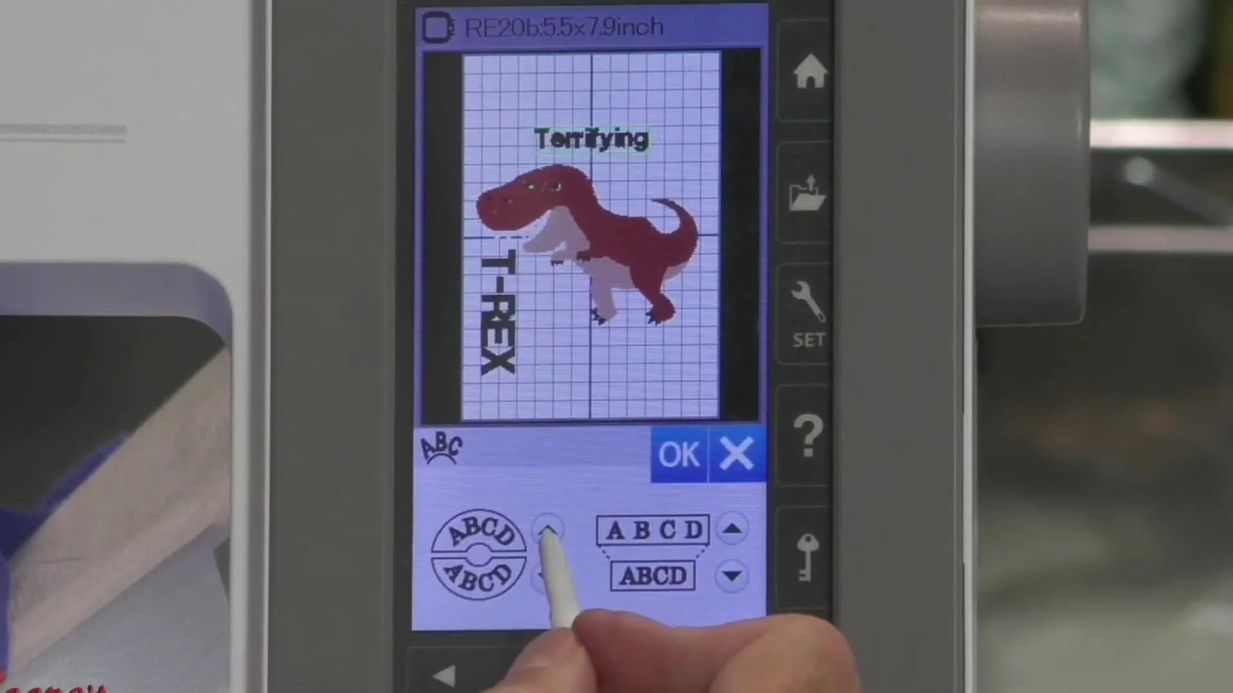 The finished T‑Rex embroidery is held up to the camera, showing the dinosaur, vertical “T‑REX” text, arced “Terrifying,” and a border element underneath.