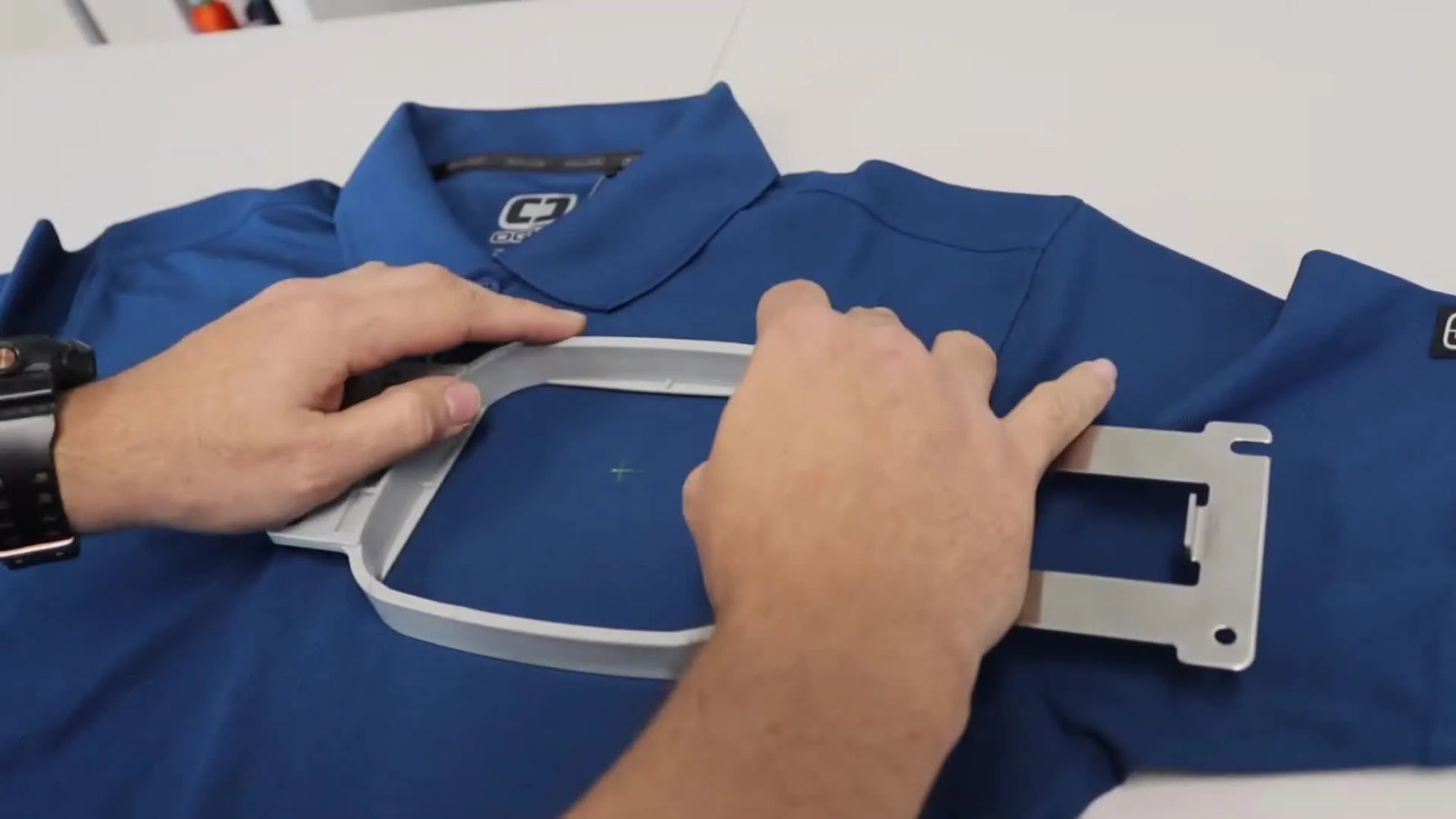 Close-up of hands pressing a tubular hoop frame into the bottom ring to secure a polo shirt for left-chest embroidery.