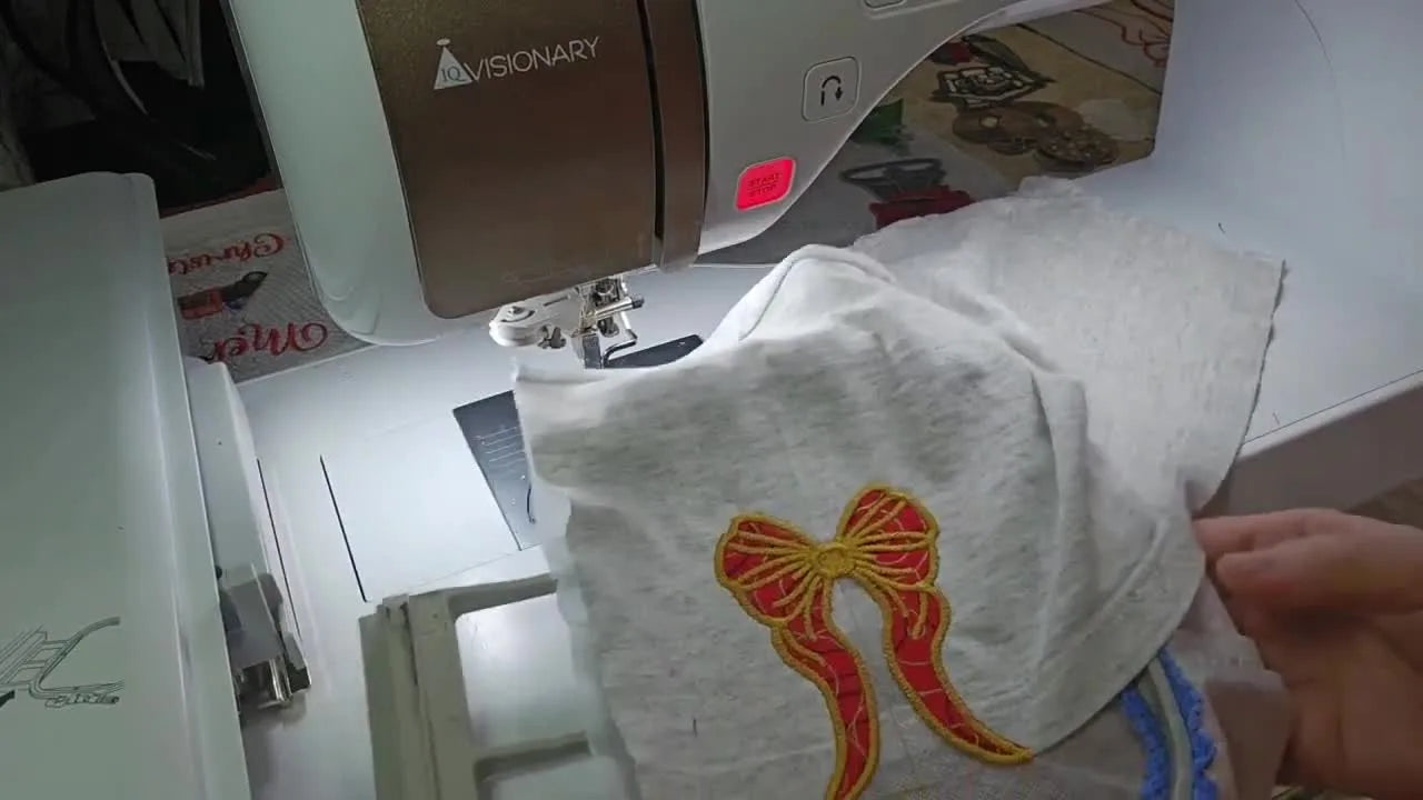 A close-up of the finished gold-outlined red bow embroidered neatly along the side seam of a heather grey child’s T-shirt.