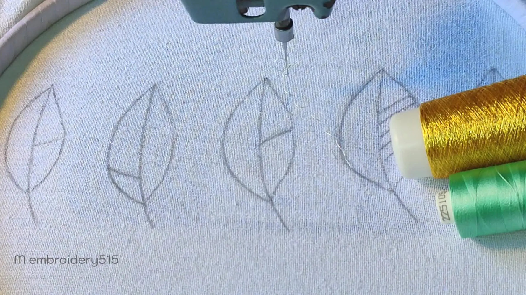 Overhead shot of white fabric in an embroidery hoop with drawn leaf outlines and two spools of embroidery thread (green and gold).