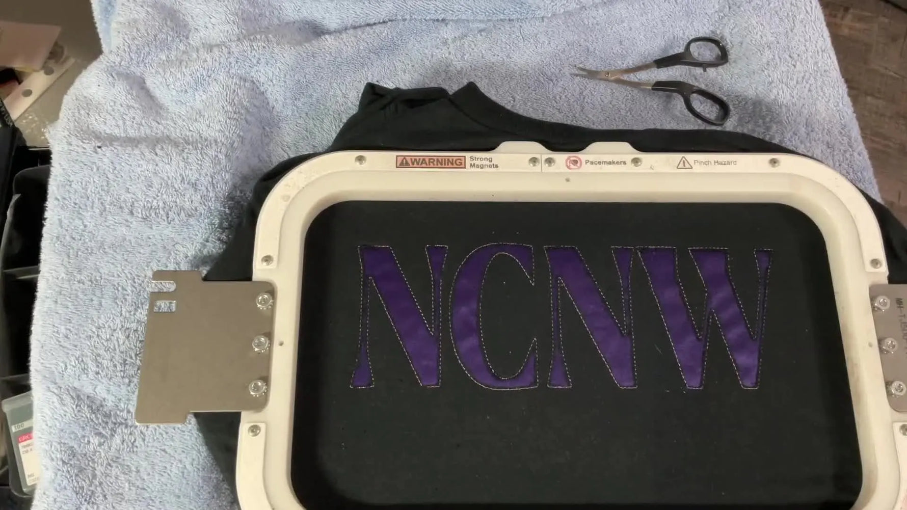 A hooped black T-shirt after the reverse-appliqué cutout reveals purple letters underneath, ready for the next stitch pass.