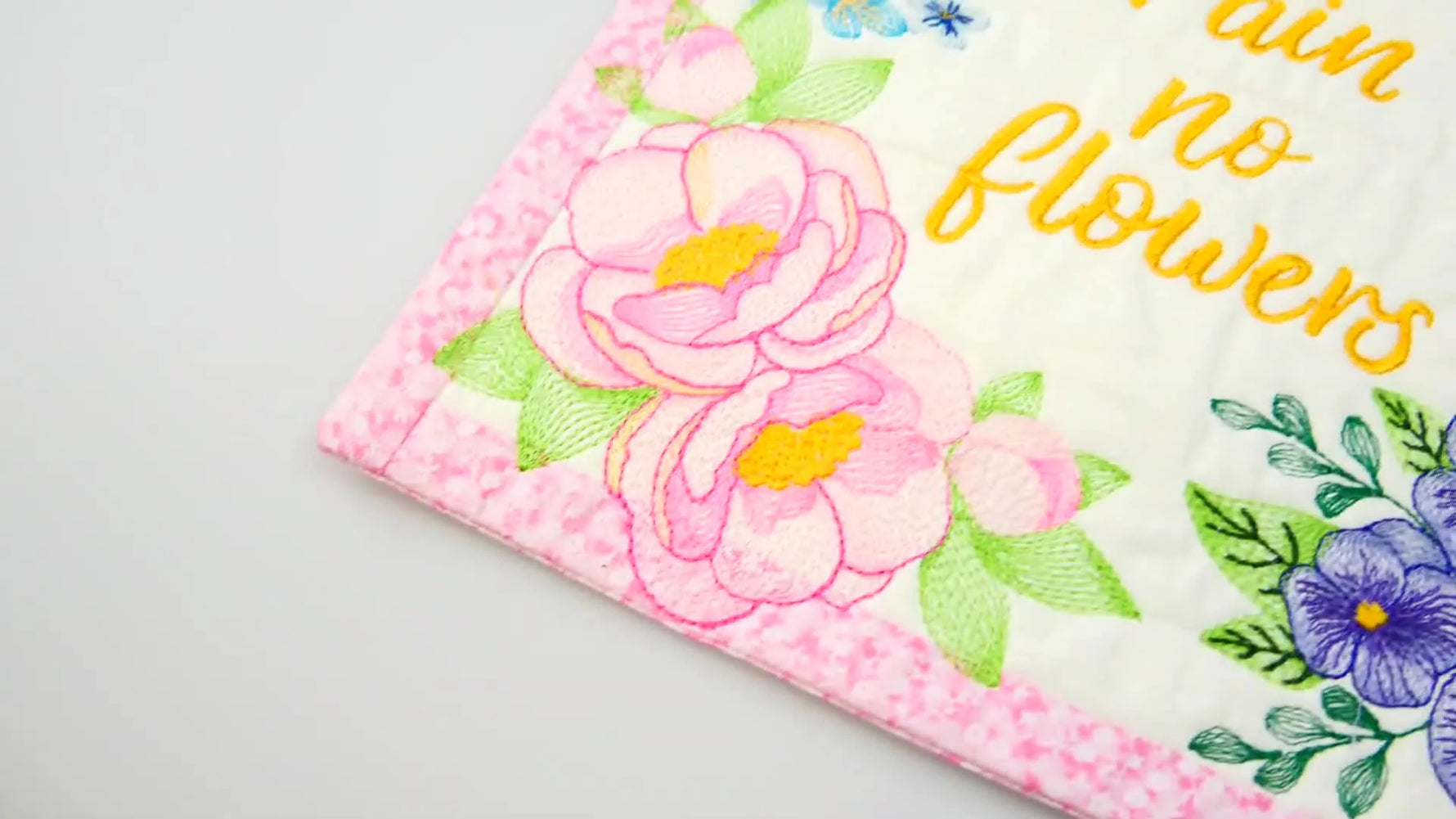 The completed “No Rain No Flowers” wall hanger lies flat, showing crisp borders, floral embroidery, and two hanging loops aligned at the top.