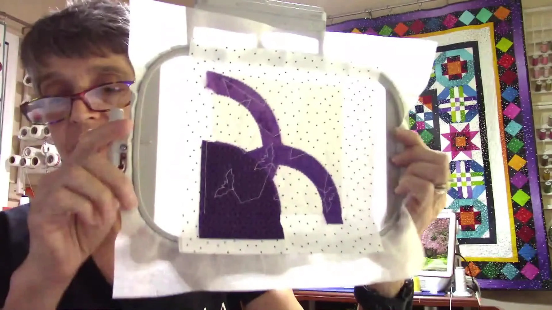 A hooped Halloween quilt block is held up close to the camera, showing a bat design stitched with glow-in-the-dark thread on purple fabric.