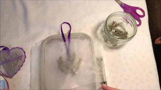 Hands tape a floating stabilizer layer to the plastic rim of a 4x4 hoop to secure an in-the-hoop lavender sachet sandwich.