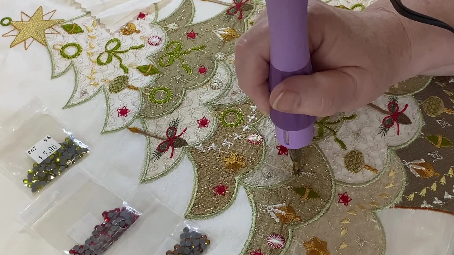 A hotfix applicator is pressed straight down onto a crystal already positioned on an embroidered Christmas tree for precise, slip-free placement.