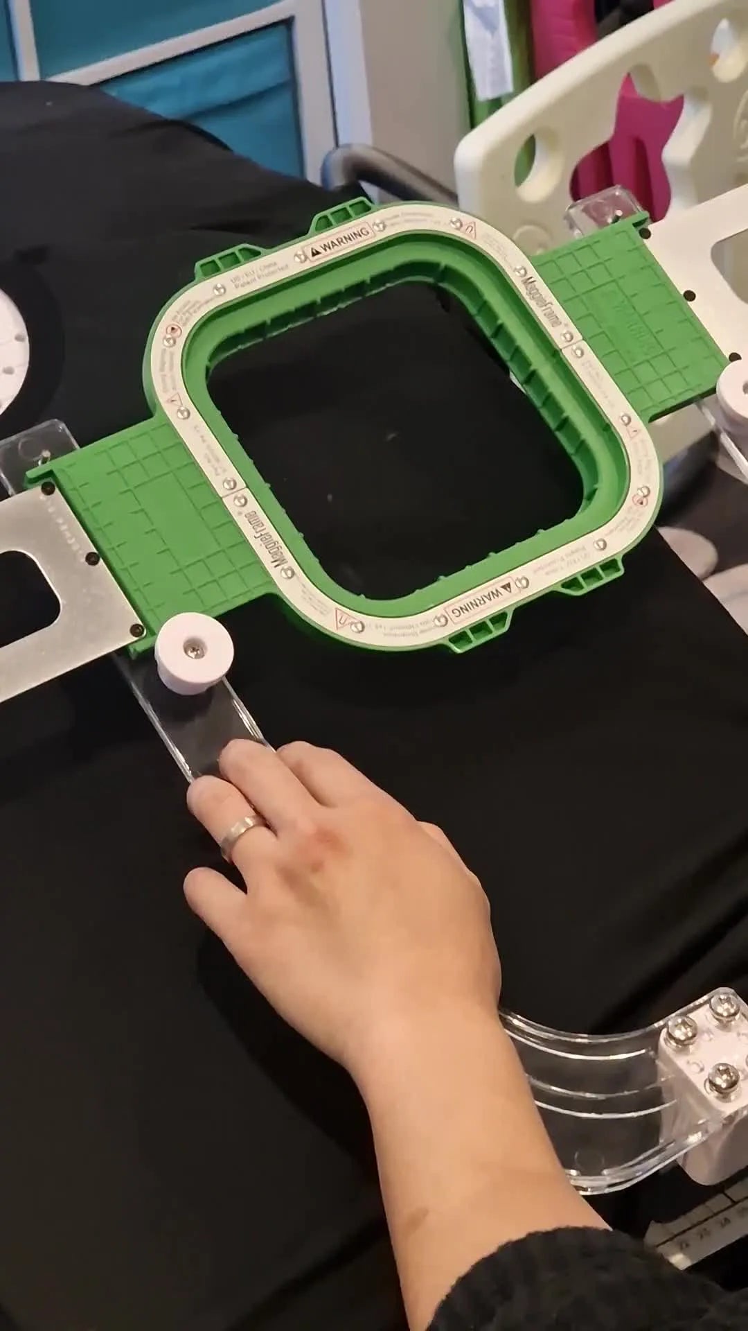 Hands press the white alignment circles to snap the MaggieFrame magnetic hoop shut on a black T-shirt in the Sew Tech hooping station.