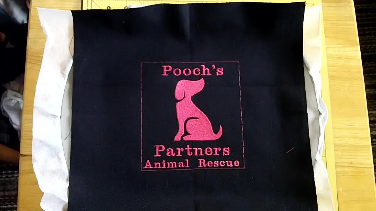 An overhead view of the finished pink dog-and-text embroidery stitched on hooped black denim with white stabilizer visible underneath.