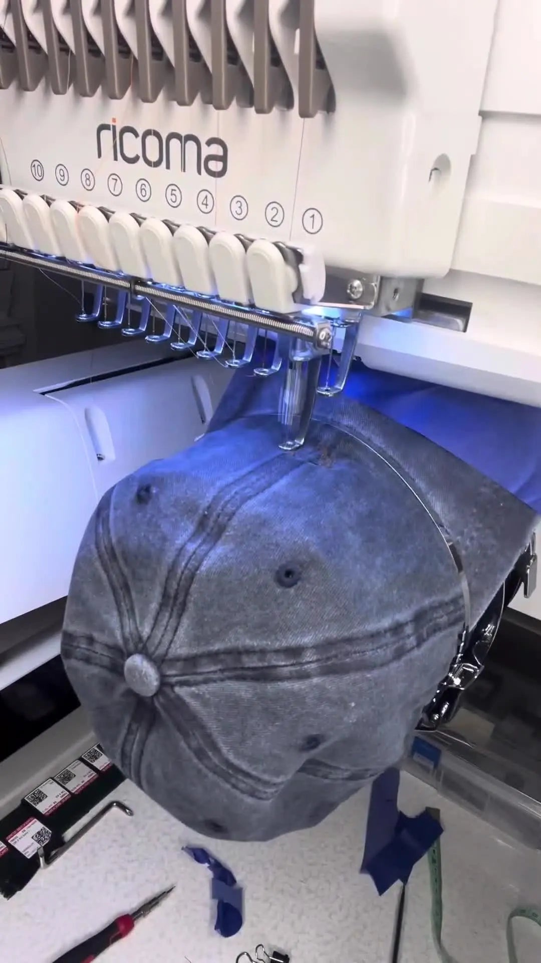 A Ricoma multi-needle machine stitches a detailed design on a soft, unstructured dad hat mounted on a cap driver.