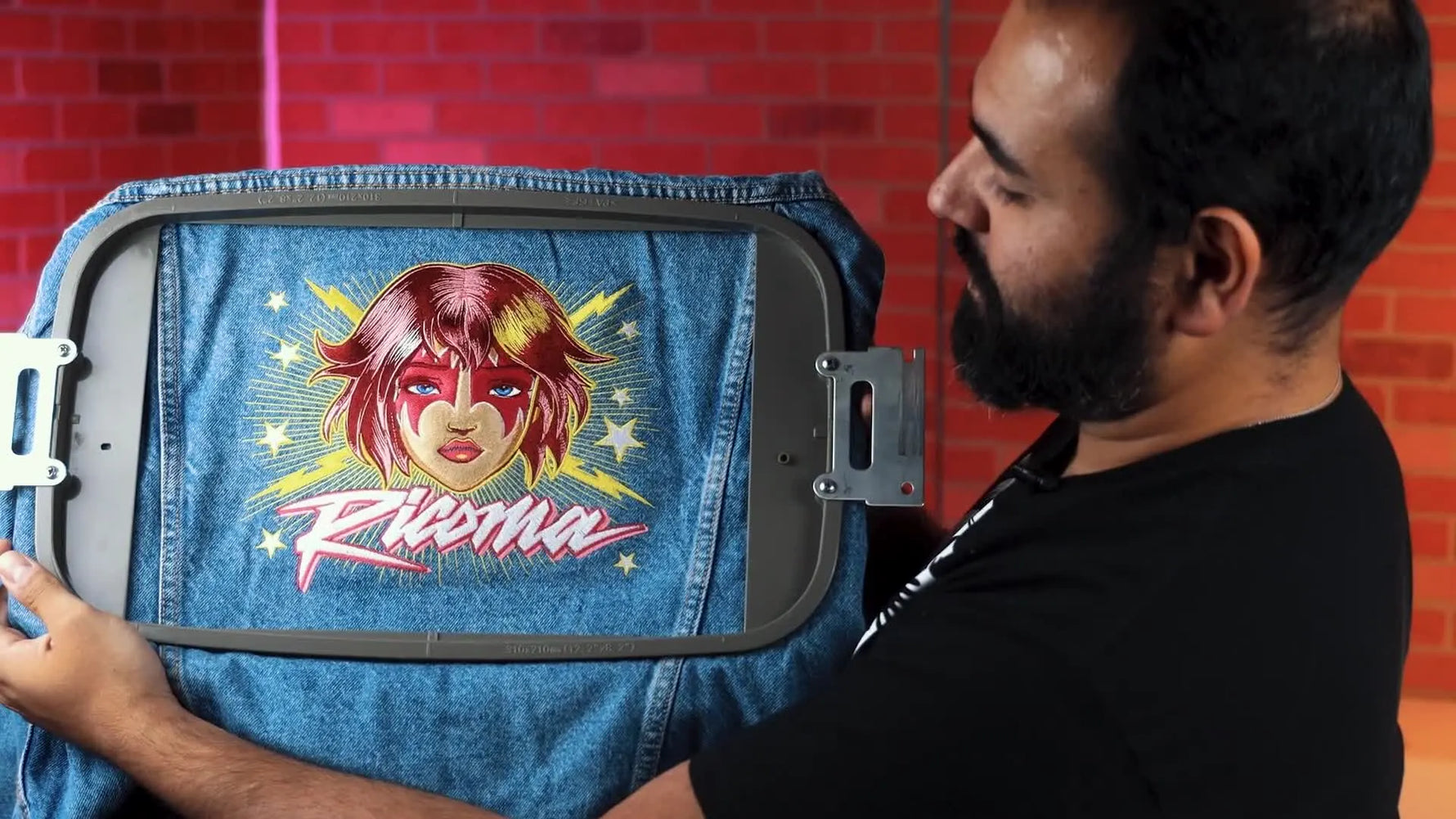 The host holds up the finished, vibrant multi-color rock design stitched on the back of a denim jacket while it’s still hooped.