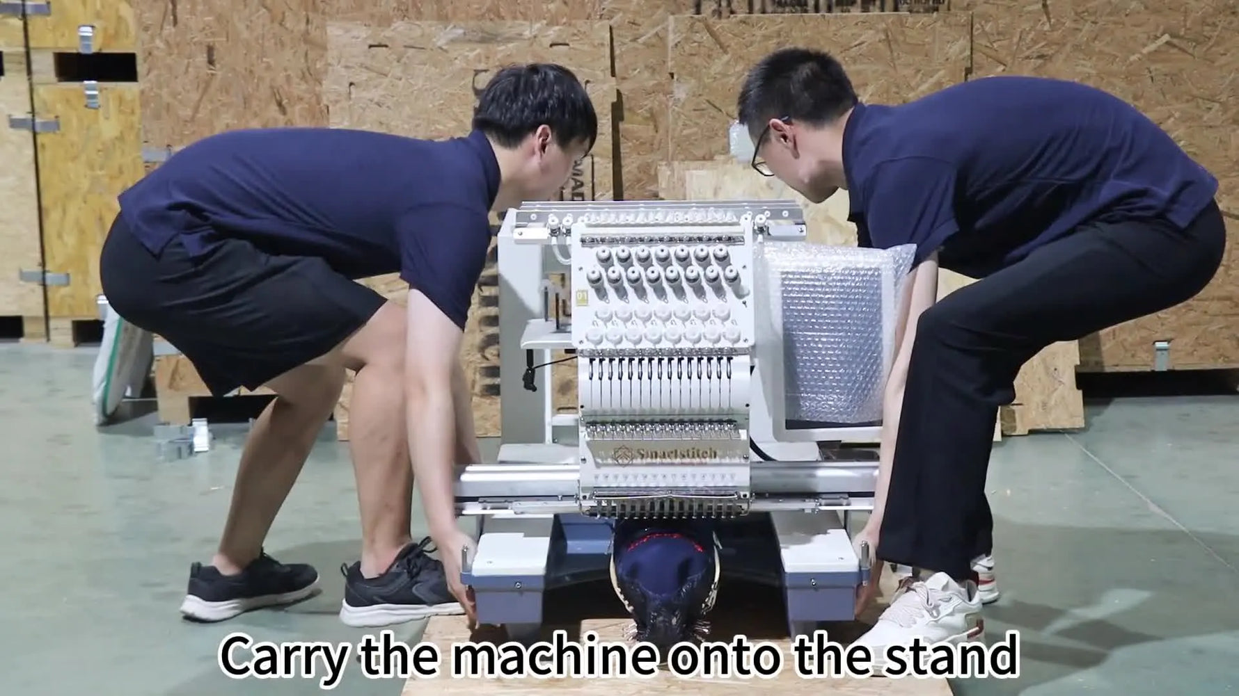 Two operators lift a commercial Smartstitch multi-needle embroidery machine onto the assembled rolling stand for final mounting.