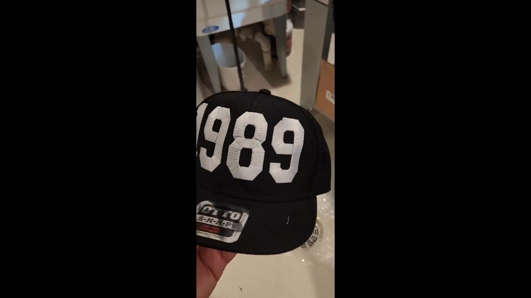 A finished black Otto snapback is held close to the camera, showing a bold white “1989” embroidery centered on the front panel.