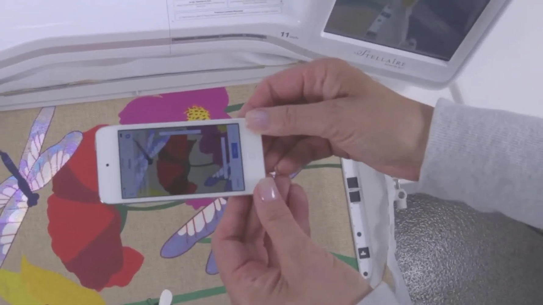 A phone is held above a hooped floral fabric while the Brother Stellaire displays the placement workflow for My Design Snap.
