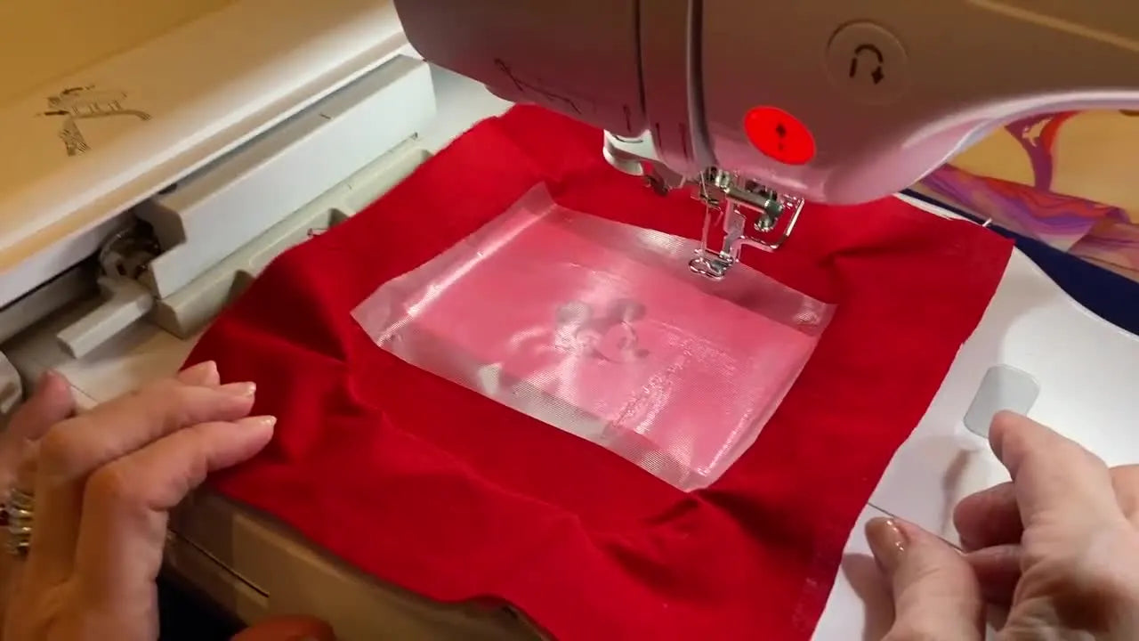 A close-up of the Brother Luminaire projecting a Mickey Mouse design onto red batik fabric in the hoop for precise placement before stitching.
