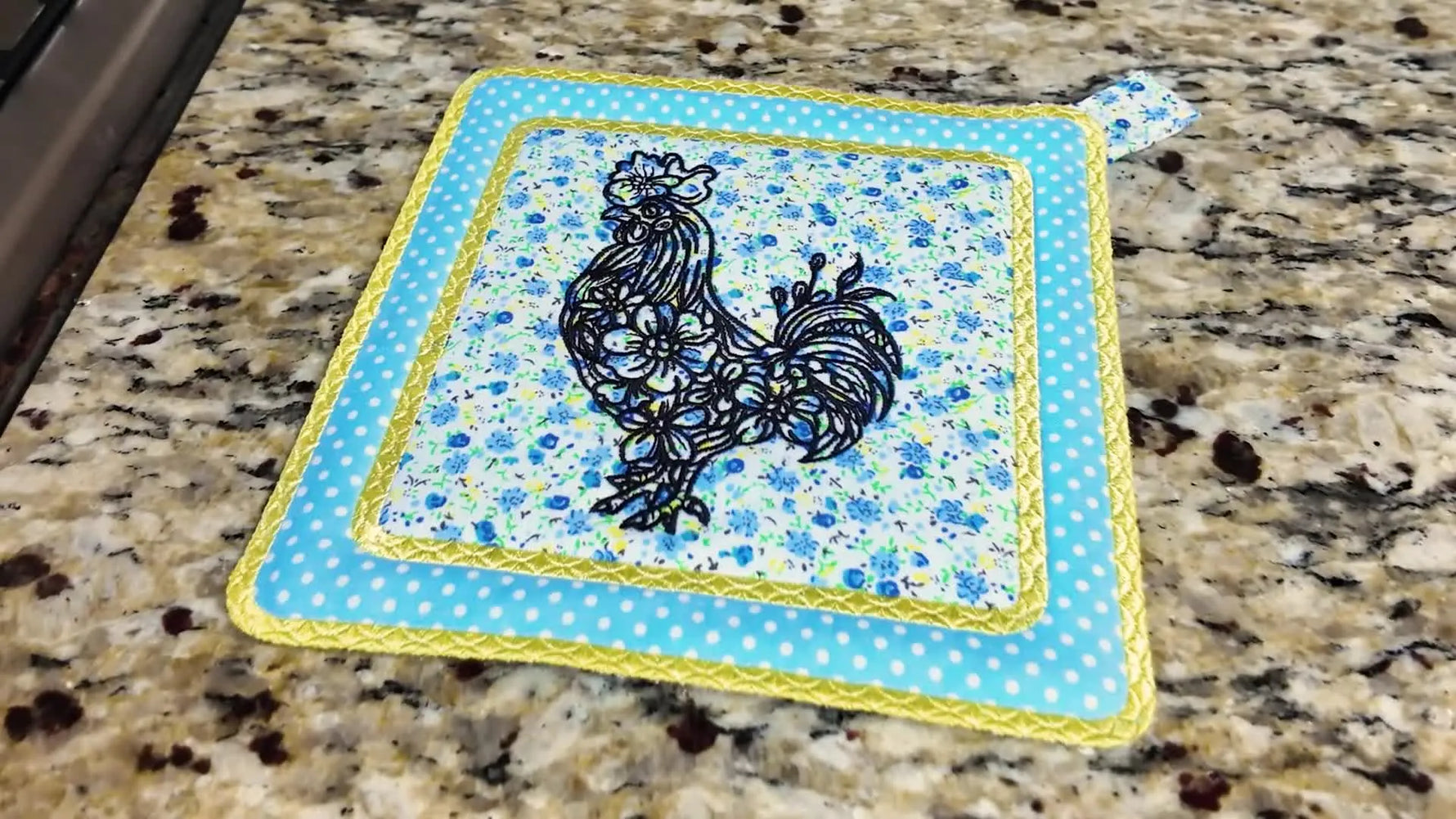 The finished rooster-themed in-the-hoop trivet sits neatly on a granite countertop with a bold satin-stitched border and a clean appliqué frame.
