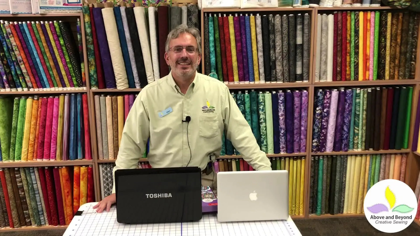 A shop owner stands behind a counter with colorful fabric bolts, demonstrating Premier+2 software with both a Toshiba laptop and a MacBook to highlight PC/Mac compatibility.