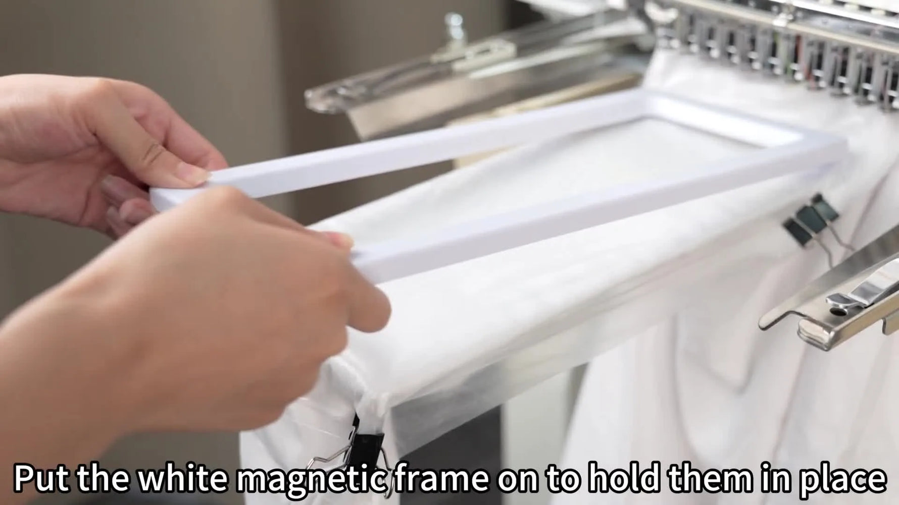 A white rectangular magnetic top frame is being placed onto a sleeve on the Smartstitch arm to lock the fabric flat before stitching.