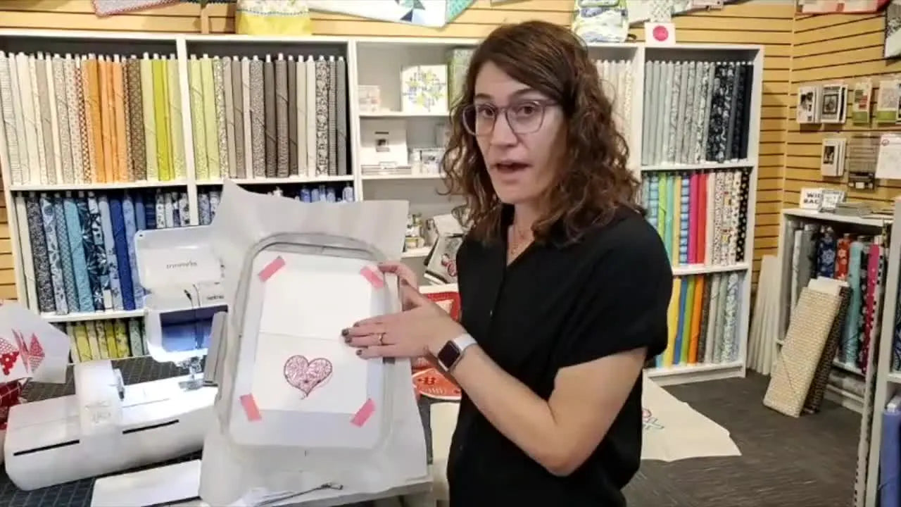 The host points to pink tape securing cardstock on top of cutaway stabilizer inside a standard embroidery hoop.
