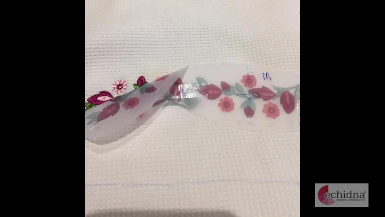 A translucent placement template is peeled back to reveal perfectly aligned embroidery underneath, confirming a clean multi-hoop match.
