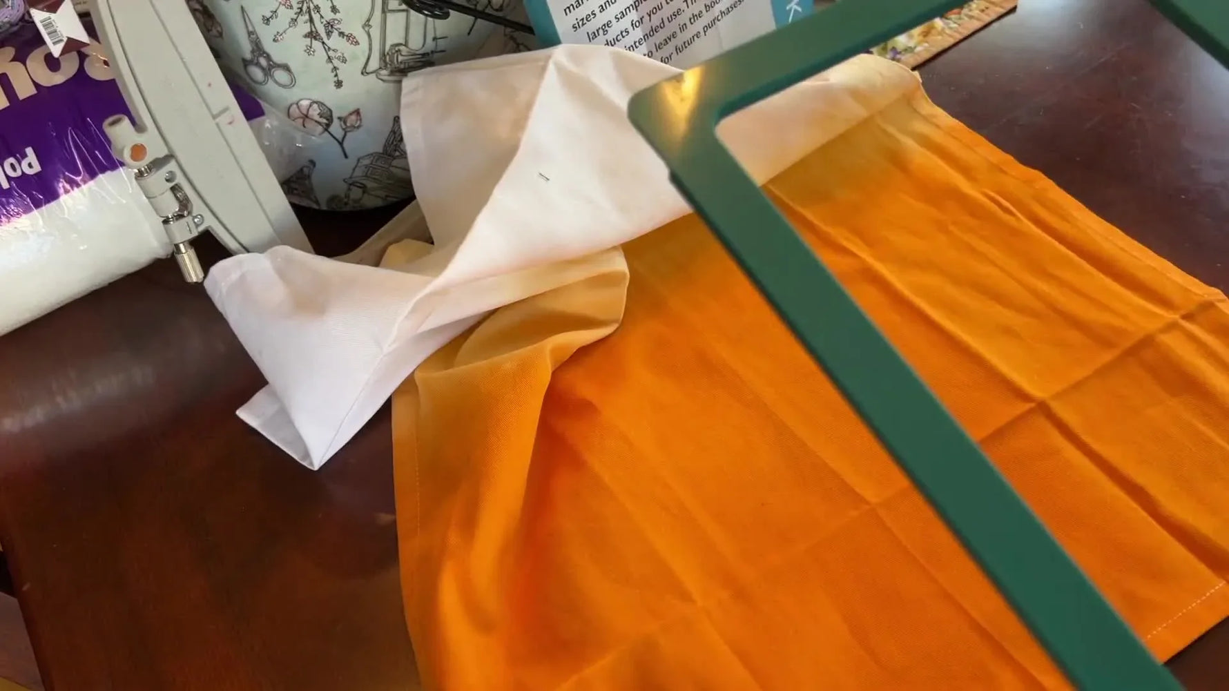 Hands snap a teal 5x7 magnetic hoop onto an orange ombré tea towel with stabilizer underneath for fast, burn-free hooping.