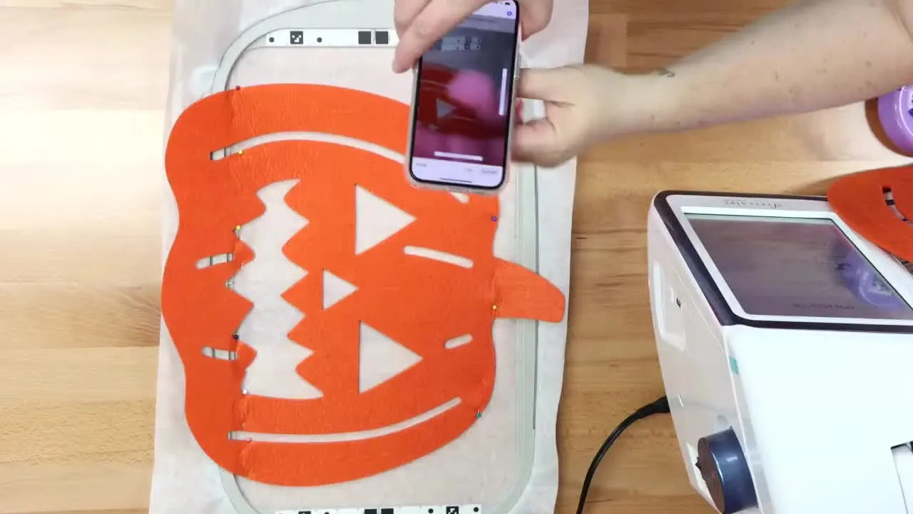 An overhead shot of a smartphone held parallel above a hooped felt placemat while My Design Snap scans the embroidery frame for perfect placement.