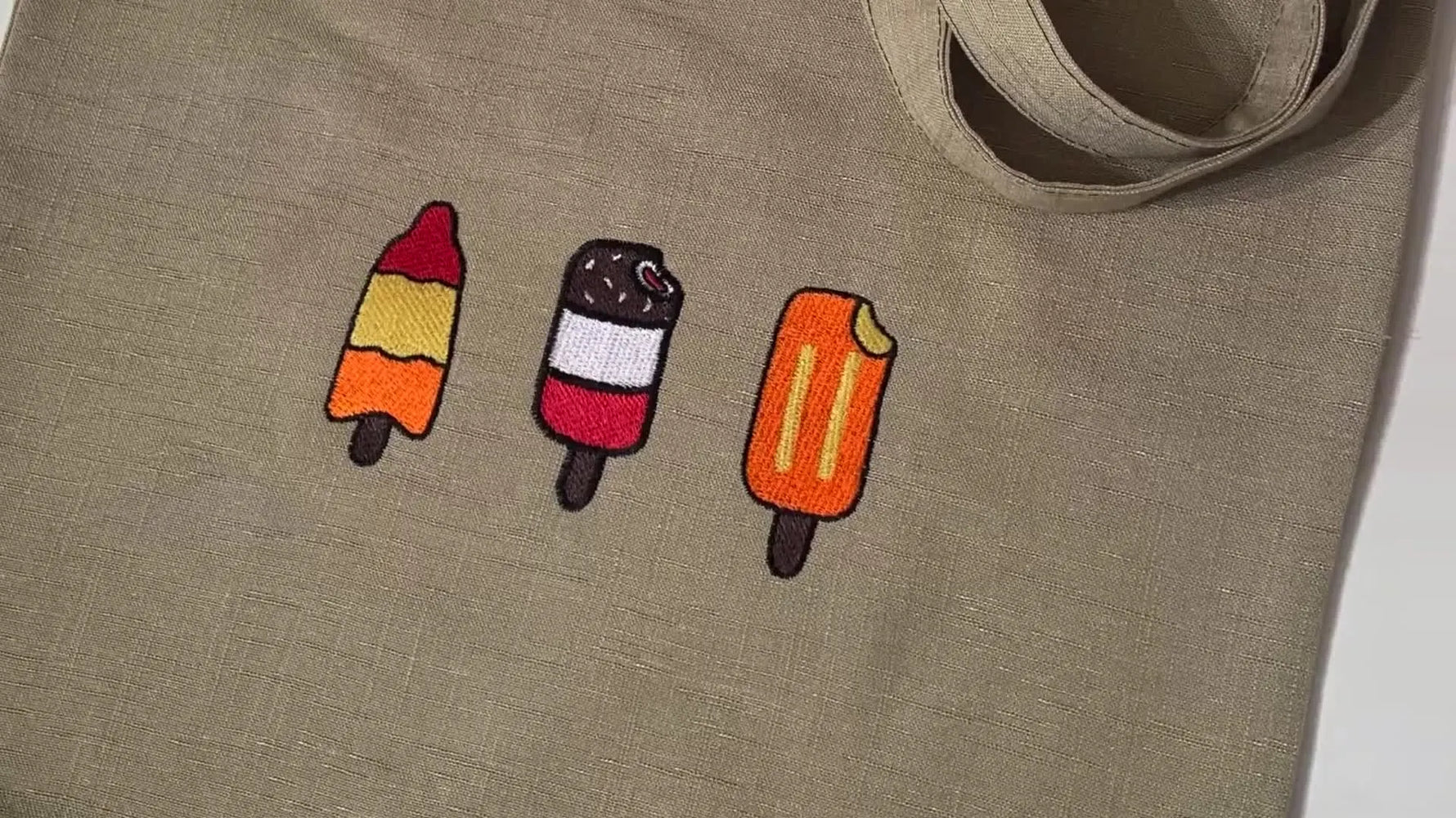 A finished oatmeal linen tote bag showing three cleanly stitched popsicle designs in a neat row.