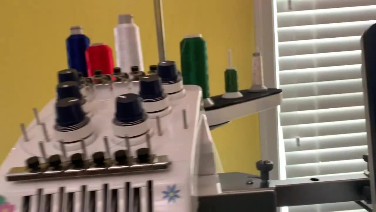 A close-up of a multi-needle embroidery machine stitching an ivory-on-ivory monogram on a hooped dinner napkin.