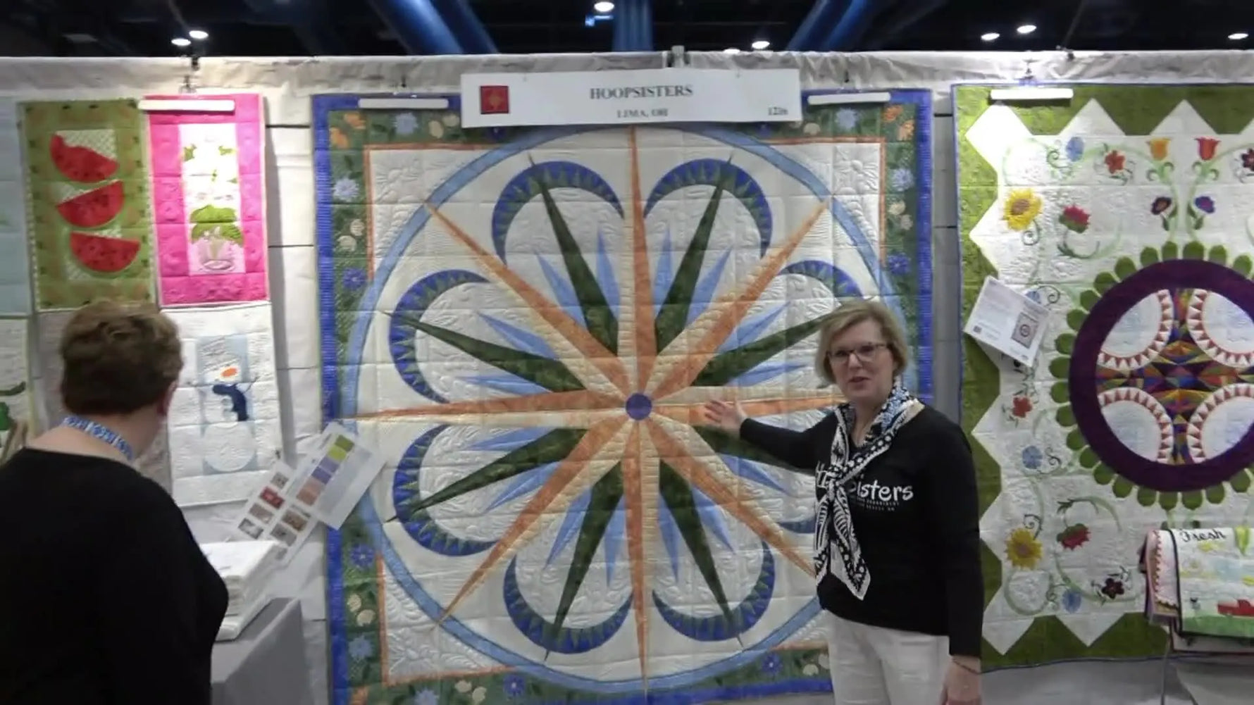Annie gestures toward the large HoopSisters “Navigator” heirloom quilt hanging on the booth wall at the 2022 Houston Quilt Market.