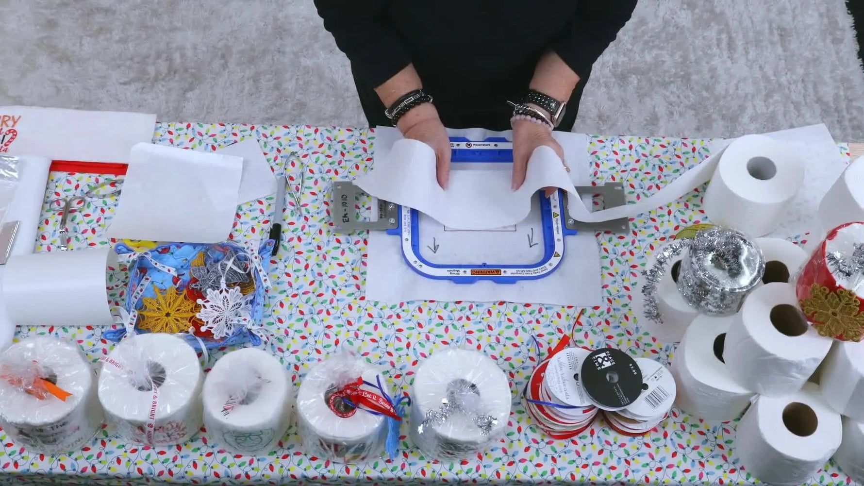 A close-up overhead shot shows a sheet of toilet paper being gently pressed into a sticky stabilizer window inside a magnetic hoop before stitching.