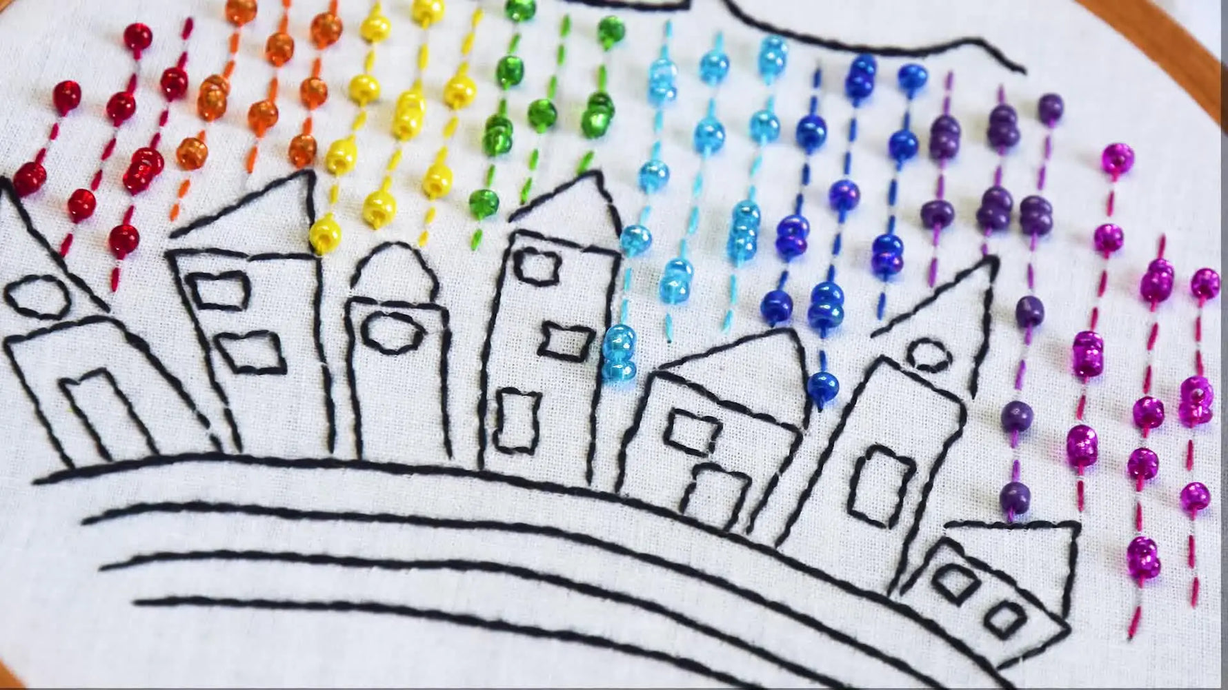 Finished embroidered piece with whimsical houses and rainbow beaded rain in an embroidery hoop.