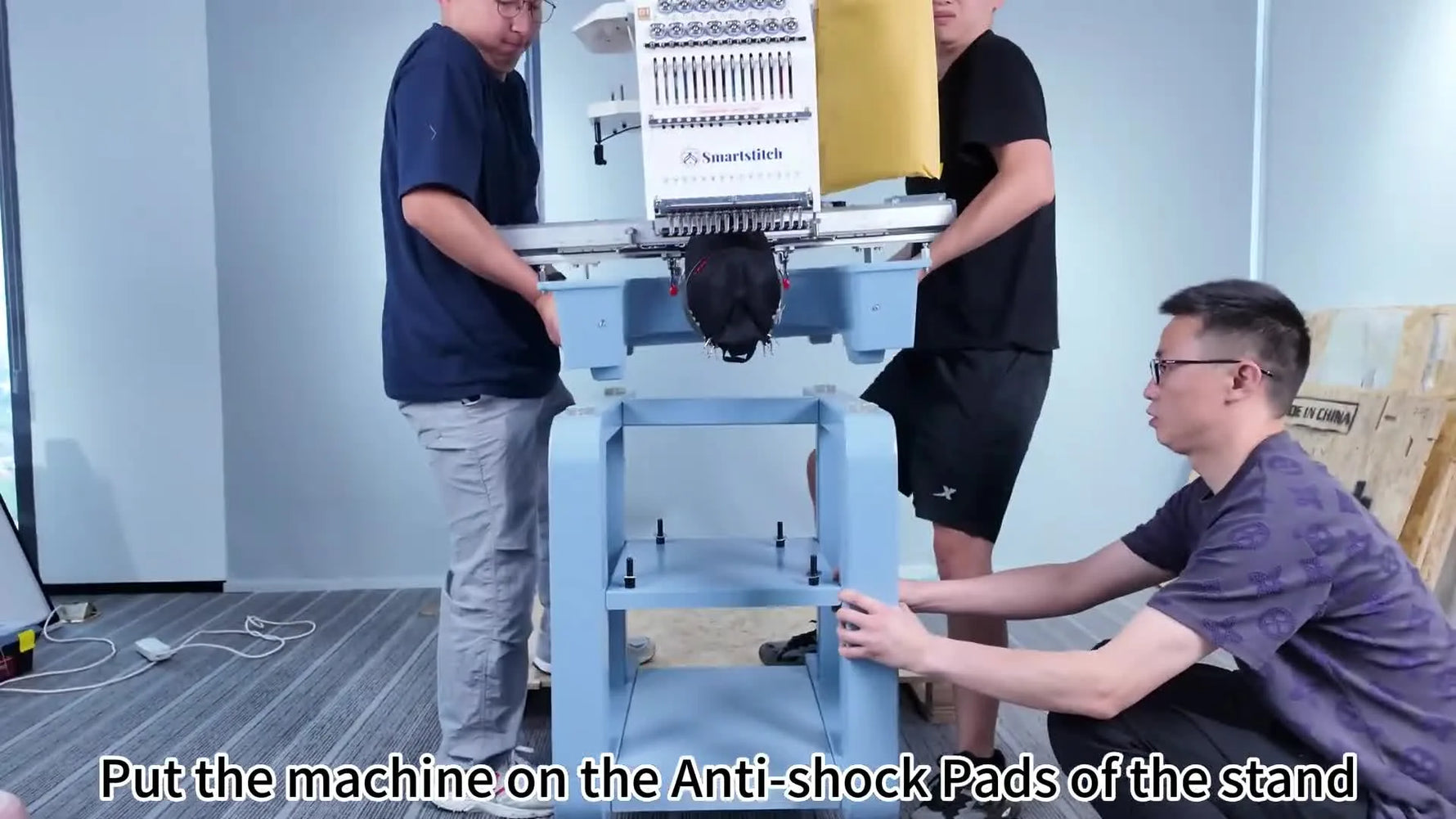 Three people lift a Smartstitch multi-needle embroidery machine onto the assembled stand, aligning it with the anti-shock pads before bolting it down.
