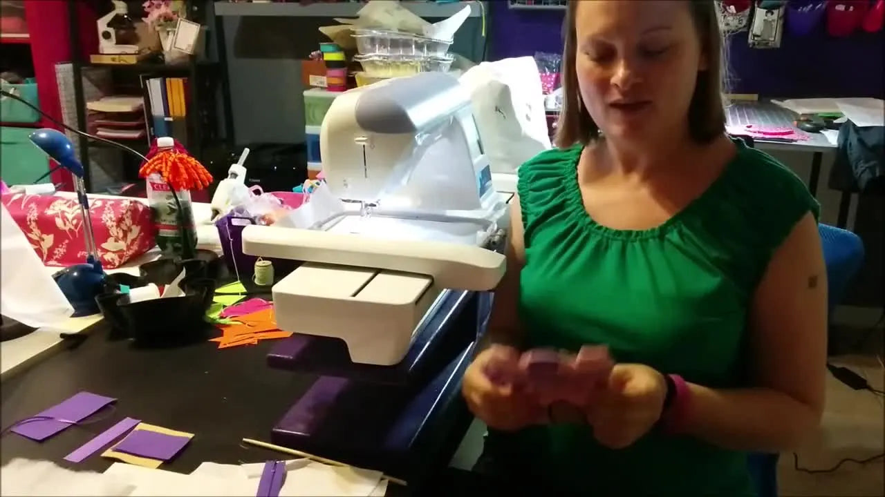 A finished pink-and-purple in-the-hoop castle zipper bag is held up in front of an embroidery machine, showing the crisp shape and matching zipper pull.