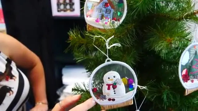 A finished snowman snow globe ornament hangs on a Christmas tree, showing a clear PVC window with loose sequins inside.